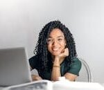 Joyful businesswoman with curly hair smiling at camera while using laptop indoors.