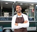 Smiling young ethnic male waiter in apron standing with arms folded near modern food truck and looking at camera contentedly